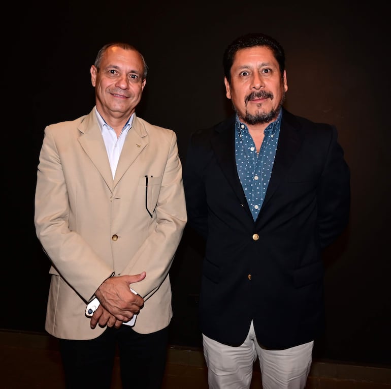 Dos hombres en primer plano, uno con saco claro y expresión amable, el otro con saco oscuro y camisa a rayas, en entorno formal.