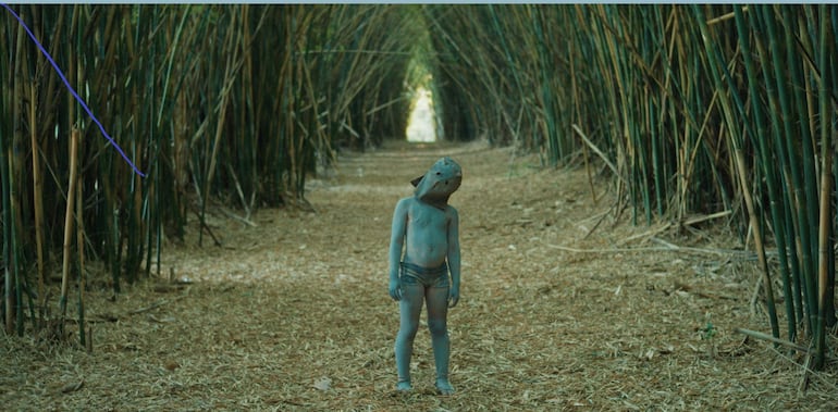 Niño en medio de cañas de bambú, cuerpo azul y máscara de pez, observa a la cámara en un ambiente boscoso.