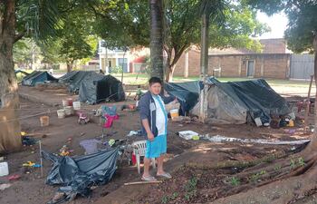 Hombre de camiseta blanca y pantalones cortos celestes de pie en campamento de carpas negras, rodeado de objetos dispersos.