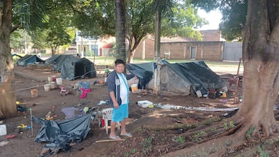 Hombre de camiseta blanca y pantalones cortos celestes de pie en campamento de carpas negras, rodeado de objetos dispersos.