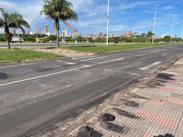 Calle desierta con pavimento irregular, palmeras a un lado y edificios al fondo bajo un cielo despejado.