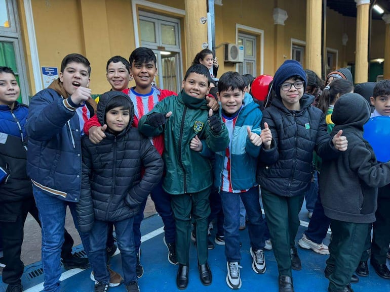 Grupo de niños sonrientes con camisetas rojas y blancas, levantando pulgares, en un entorno festivo y frío.