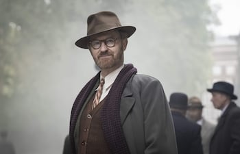 Mark Gatiss con sombrero y abrigo vintage, rodeado de personas similares en un entorno urbano y neblinoso.
