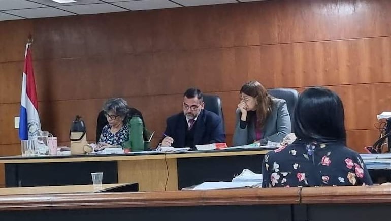 Andrea Vera, con cabello rizado y anteojos, revisa documentos mientras un hombre escribe y otra mujer observa en un ambiente judicial.