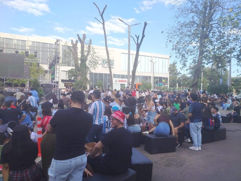 Multitud de personas con camisetas azul y blancas animan en la Embajada de Argentina, al frente una pantalla que transmite un partido.