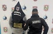 Hombre con capucha y chaleco de Interpol se encuentra de espaldas, acompañado por otro con abrigo negro, en un ambiente interior con logotipos de Interpol.