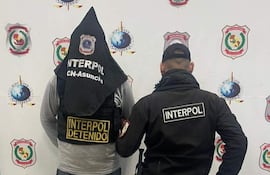 Hombre con capucha y chaleco de Interpol se encuentra de espaldas, acompañado por otro con abrigo negro, en un ambiente interior con logotipos de Interpol.