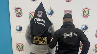Hombre con capucha y chaleco de Interpol se encuentra de espaldas, acompañado por otro con abrigo negro, en un ambiente interior con logotipos de Interpol.