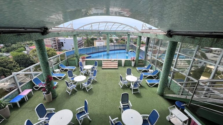 Área exterior con sillas y mesas blancas en un ambiente urbano, piscina al fondo y vegetación alrededor.