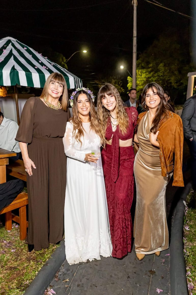 Pelusa Rubin, novia vestida de blanco, sonríe rodeada de tres mujeres en vestido burdeos, dorado y marrón, en ambiente festivo.