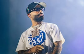 Arcángel en escenario vistiendo camiseta blanca y gorra oscura, con cadena prominente, actuando en un evento musical.