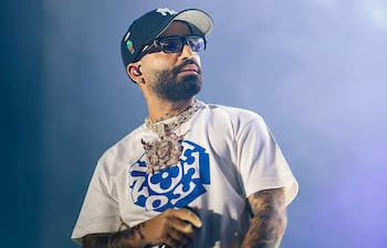 Arcángel en escenario vistiendo camiseta blanca y gorra oscura, con cadena prominente, actuando en un evento musical.