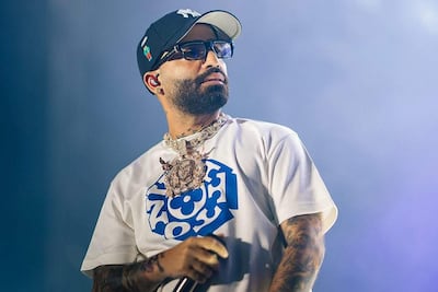 Arcángel en escenario vistiendo camiseta blanca y gorra oscura, con cadena prominente, actuando en un evento musical.
