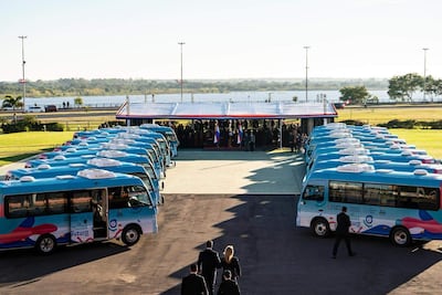 Multitud observa la presentación de minibuses azules bajo una estructura cubierta, rodeada de vegetación y un río al fondo.
