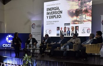 Seis panelistas en el conversatorio sobre energía, inversión y empleo, atentos en su atuendo formal, rodeados de un ambiente ordenado y banners informativos.