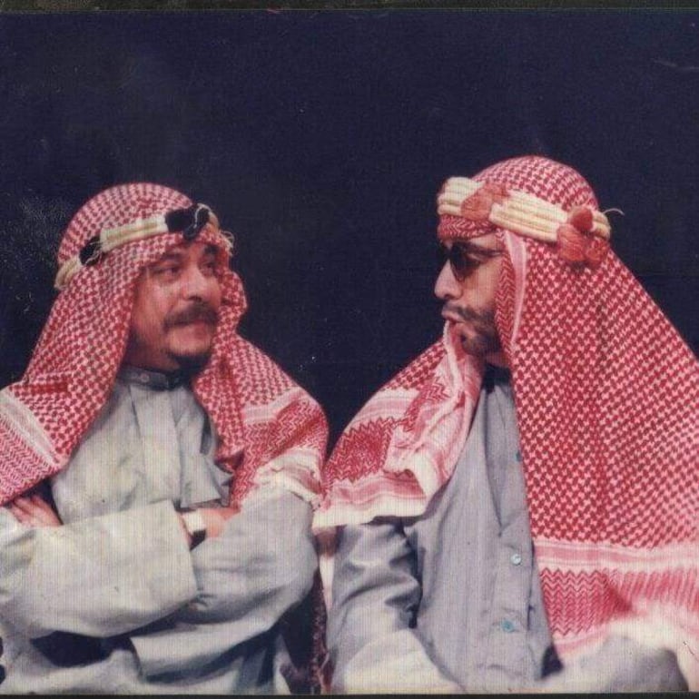 Dos hombres con vestimenta árabe, uno sonríe y el otro observa serio en un entorno oscuro.
