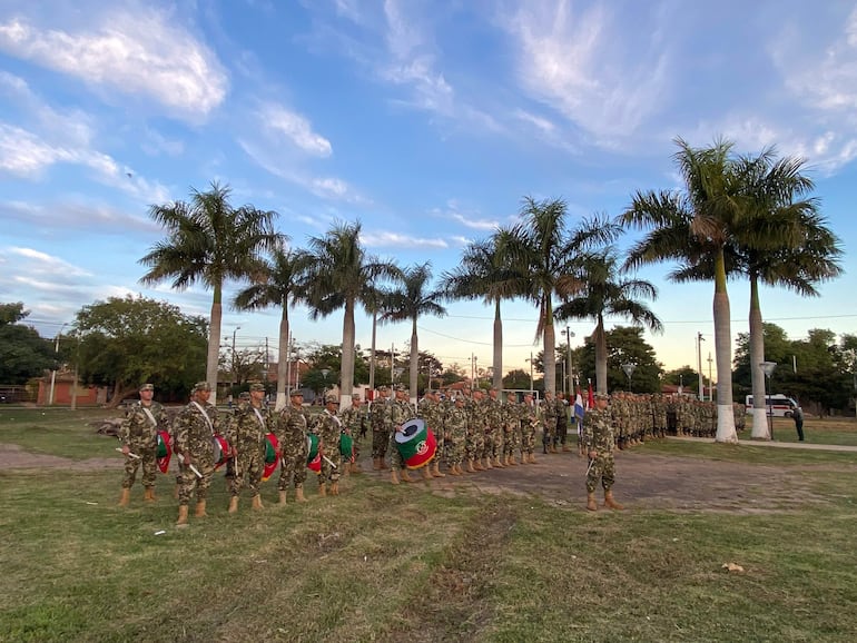 Soldados en formación sostienen banderas paraguayas, vestidos con uniformes camuflados, rodeados de palmeras y césped verde.