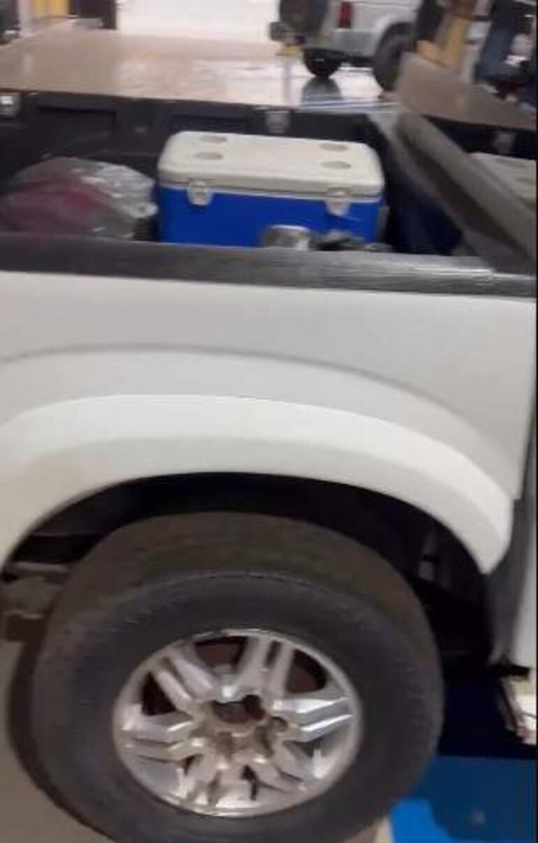 Camioneta blanca con rueda de repuesto y cooler azul en un taller, sin personas visibles en la imagen.