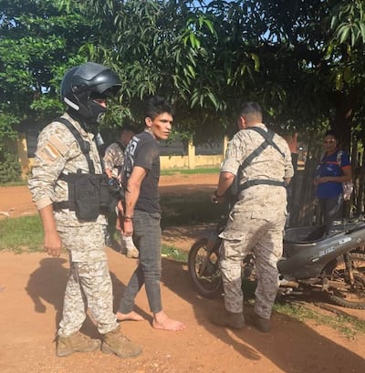 Hombre descalzo y con camiseta negra bajo custodia de militares en camuflado, rodeado de árboles y una motocicleta.