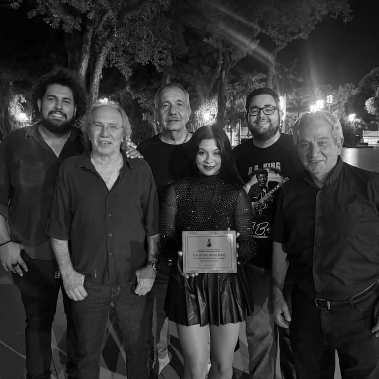 Mujer en el centro con premio, rodeada de cinco personas sonrientes en ambiente nocturno con luces y árboles.