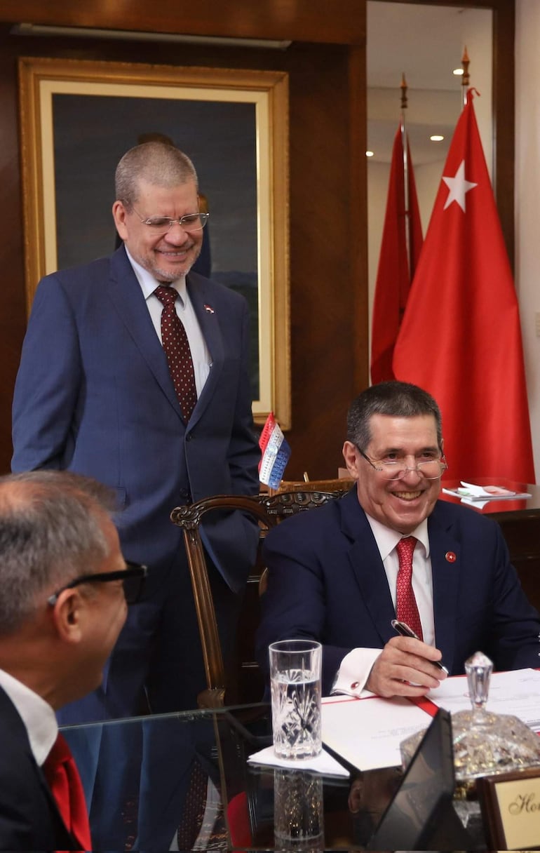 Horacio Cartes sonriente y vestido con traje azul firma documentos en un despacho, mientras Barrios Fernández lo observa.