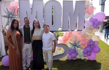 Mujer en vestido marrón y chal, hombre en chaqueta clara, mujer en blusa blanca y niño con camiseta blanca sonríen ante un letrero 'MOM'.