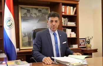 Víctor Ríos Ojeda vestido con traje oscuro y corbata azul, sentado en su oficina con expresión tranquila, junto a la bandera de Paraguay.