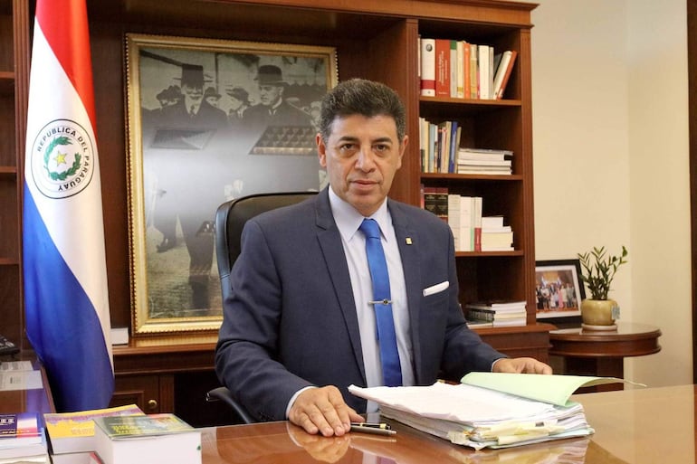 Víctor Ríos Ojeda vestido con traje oscuro y corbata azul, sentado en su oficina con expresión tranquila, junto a la bandera de Paraguay.