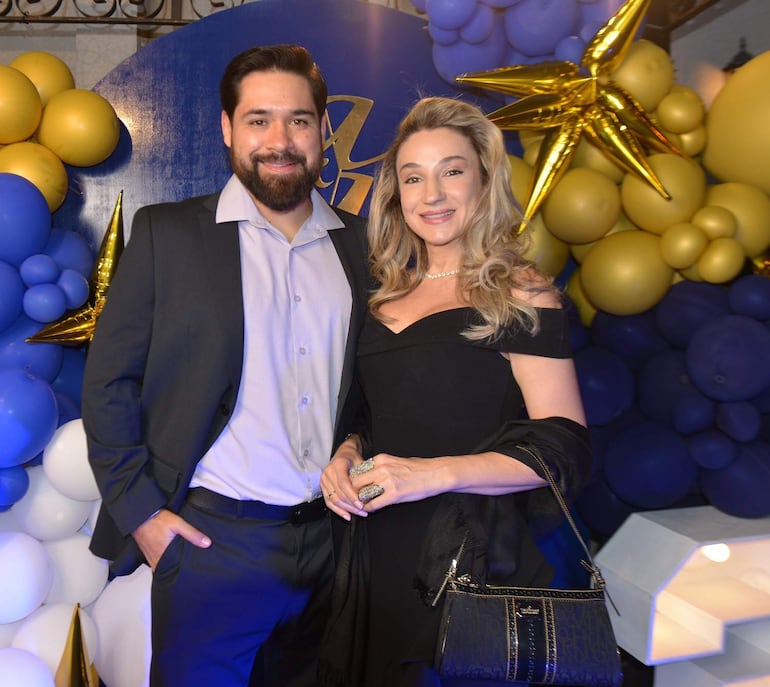 Mujer con vestido negro y chal negro sonríe junto a hombre con camisa clara y abrigo oscuro, en un salón decorado con globos.