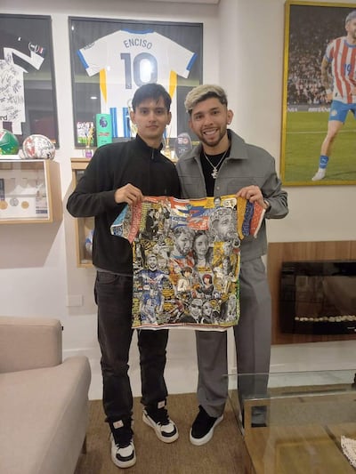 Ramón Sosa con chaqueta gris y Julio Enciso en sudadera negra sosteniendo camiseta colorida, sonriendo en interior decorado con trofeos.