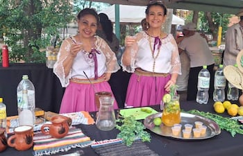 Mujeres en trajes tradicionales con faldas rosas y blusas de encaje, sosteniendo un vaso y una jarra en un ambiente festivo con decoraciones culturales.