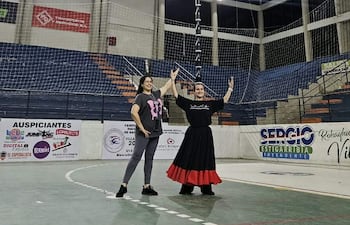 Sabrina Sosa sonríe vestida con camiseta negra y falda roja, mientras posan en una cancha de balonmano con gestos de alegría.