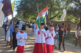 Niños en túnicas blancas y rojas sostienen candelas y banderas, rodeados de adultos en un ambiente festivo al aire libre.