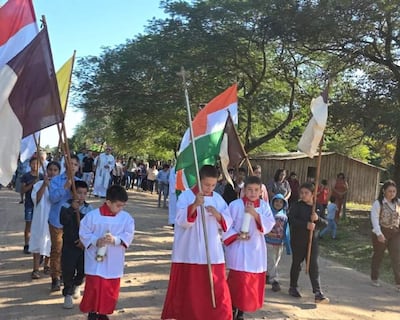 Niños en túnicas blancas y rojas sostienen candelas y banderas, rodeados de adultos en un ambiente festivo al aire libre.