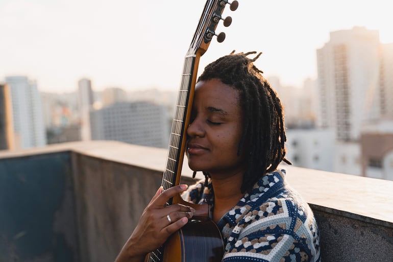 Gabriele Leite con cabello rizado y trenzado, abrazando una guitarra en un ambiente urbano al atardecer.