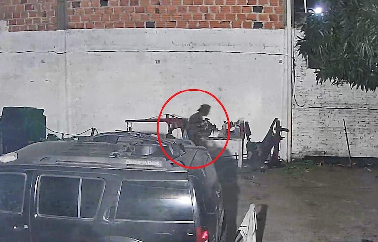 Figura humana en movimiento junto a un vehículo oscuro y una mesa desordenada, en un ambiente exterior iluminado tenuemente.