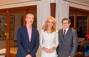 Begoña Puig en vestido blanco, flanqueada por Javier Parrondo y otro hombre con traje oscuro, en un ambiente elegante y iluminado.