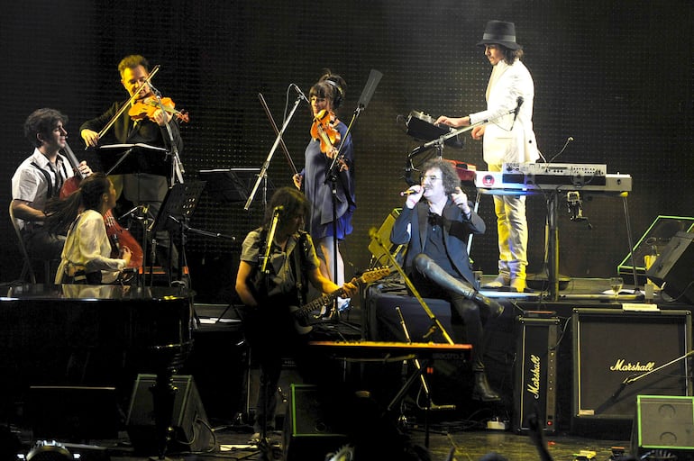 Seis músicos en escenario: hombre con violonchelo, mujer con cabello largo, cantante con chaqueta oscura y tecladista con sombrero negro.