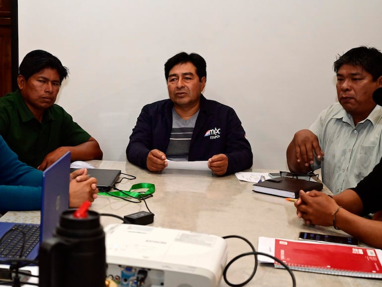 Cinco hombres sentados alrededor de una mesa con documentos y un laptop, algunos gesticulando y escuchando atentamente.