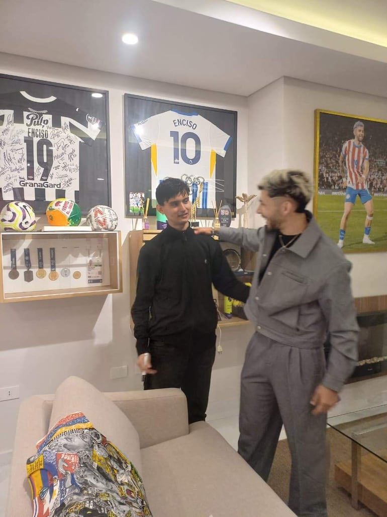 Julio Enciso con chaqueta negra escucha a Ramón Sosa en traje gris, rodeados de camisetas y balones de fútbol en un salón.