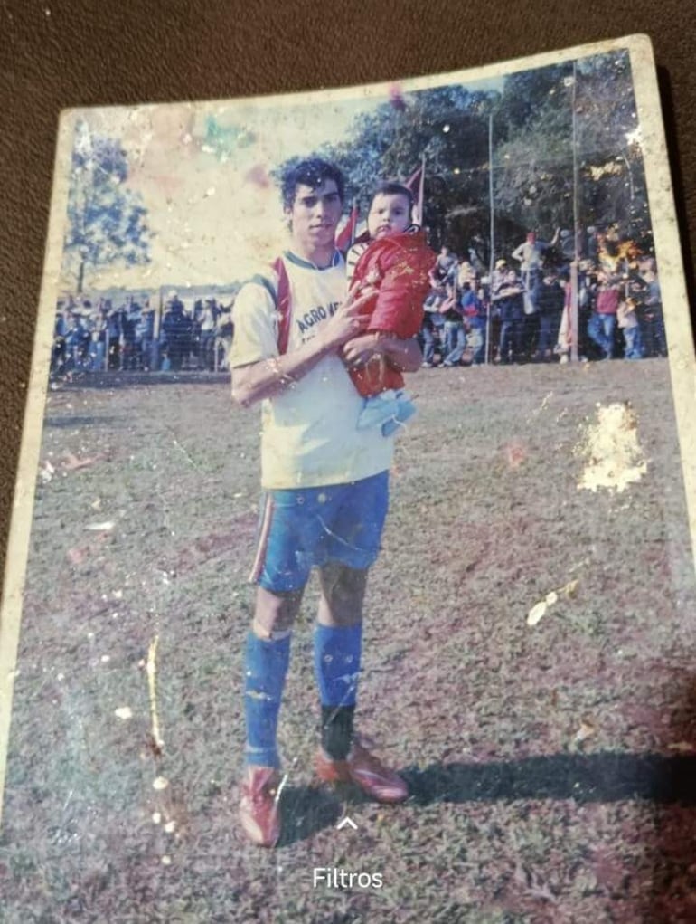 Gabriel Ávalos con camiseta blanca y detalles rojos y azules sostiene a su hermanito, ambos rodeados de espectadores y banderas.