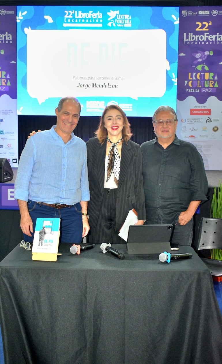 Jorge Mendelzon sonríe en el centro del escenario, mientras dos personas lo acompañan en la presentación de su libro, con decoración colorida alrededor.