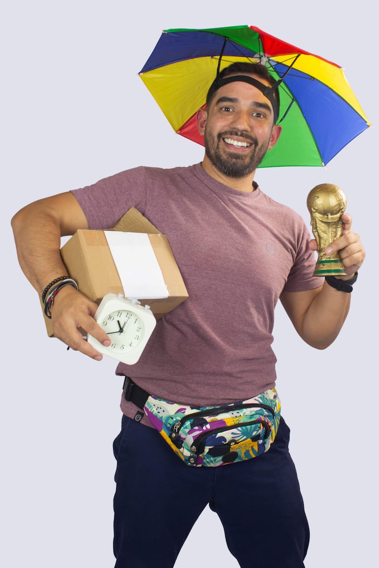 Hombre sonriente con camiseta púrpura y sombrero multicolor, sosteniendo un trofeo dorado y un reloj de alarma.
