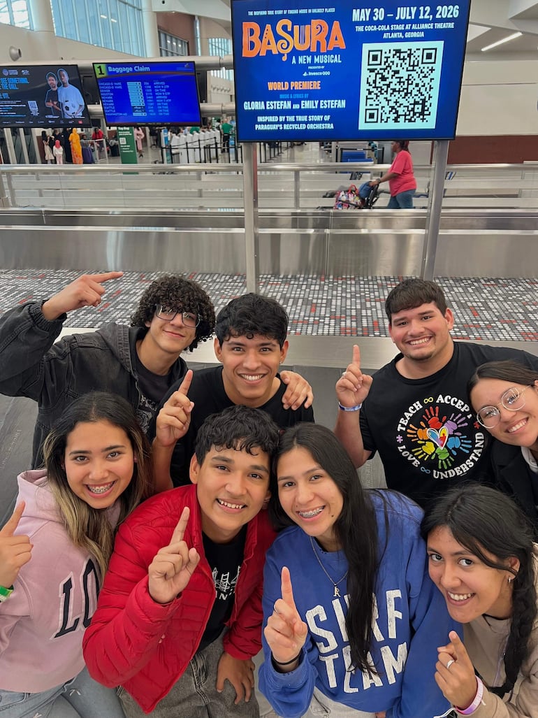 Ocho jóvenes sonrientes en un aeropuerto, levantando el dedo índice, rodeados de un ambiente festivo y una pantalla que anuncia 'Basura'.