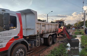 Camión rojo y blanco de la municipalidad en limpieza de calle, excavador removiendo escombros con pala bajo luz de la tarde.
