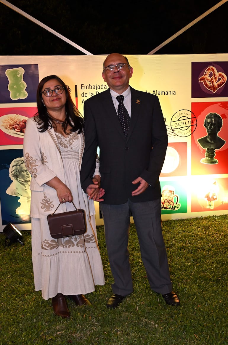 Mujer con atuendo blanco y bolso marrón sonríe junto a un hombre en chaqueta gris y corbata, en un ambiente festivo.