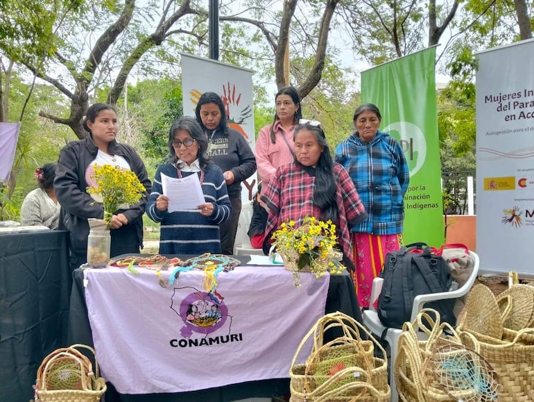 Seis mujeres indígenas en evento al aire libre, una lee documento mientras sostiene flores amarillas. Expresiones serias y mesa con artesanías.