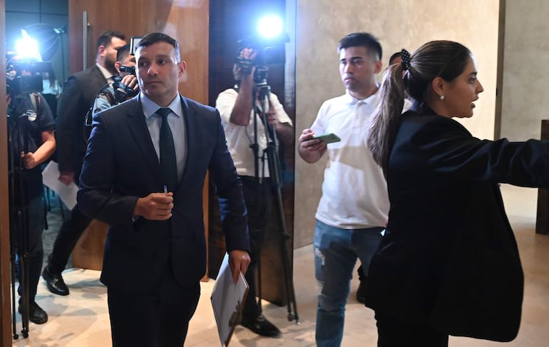 Hombre en traje oscuro con carpeta, mujer hablando, dos hombres con cámara en un ambiente formal.