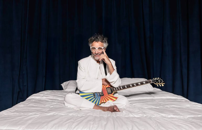 Hombre con atuendo blanco y gafas grandes, sentado en cama blanca, contemplando mientras sostiene una guitarra eléctrica colorida.