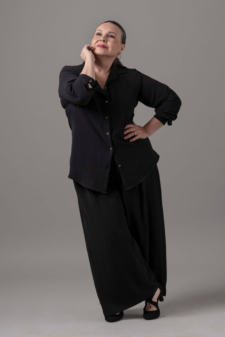 Lizza Bogado en blusa y pantalones negros, gestos pensativos, en un fondo gris en un estudio.
